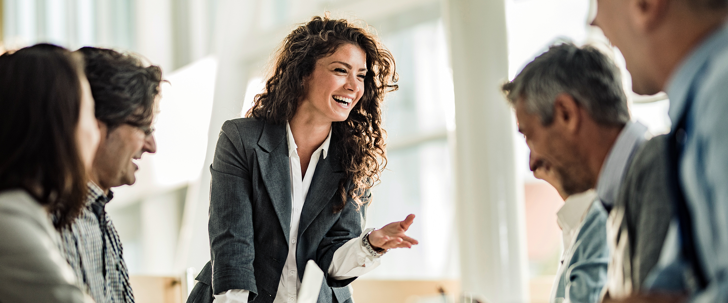 A woman standing up at a table speaking, with coworkers sitting down next to her listening and engaging.